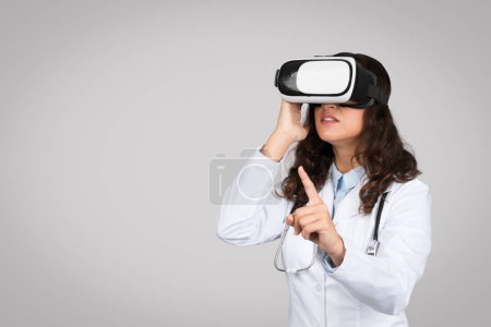 Téléchargez les photos : Jeune femme médecin en manteau blanc et lunettes vr, contrôle écran virtuel avec doigt, debout sur fond de studio gris. Sciences et technologie, médecine, soins de santé - en image libre de droit