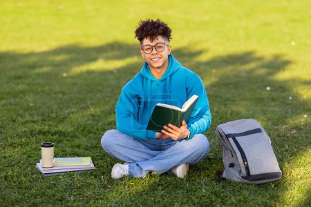 Téléchargez les photos : Pique-nique éducatif. Joyeux étudiant noir dans les lunettes de lecture livre assis sur l'herbe verte, apprendre avec café en plein air au parc, souriant à la caméra. Apprenti étudiant sur pelouse le week-end - en image libre de droit