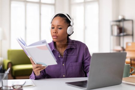 Photo for Concentrated black female student with headphones reviewing study materials and notes from textbook while simultaneously attending an online class on her laptop - Royalty Free Image