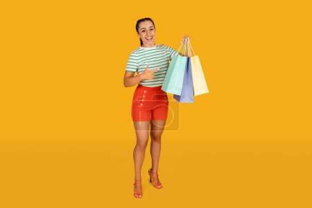 Photo for Pretty young hispanic woman pointing at colorful shopping bags in her hand and grimacing. Pretty lady enjoying season sale, showing her purchases, isolated on yellow studio , copy space - Royalty Free Image