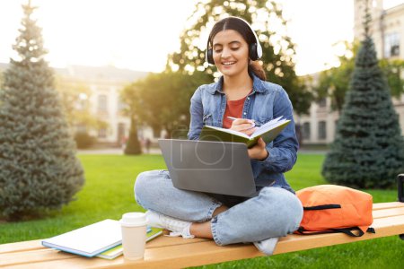 Photo for Online education, distance learning concept. Young indian woman student have video call with teacher, attending webinar, sitting on bench at park, using laptop and headset, taking notes, sun flare - Royalty Free Image