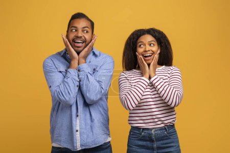 Amazed young black couple with hands on cheeks showing surprised expressions, cheerful african american man and woman standing wide-eyed and excited against yellow studio background, copy space