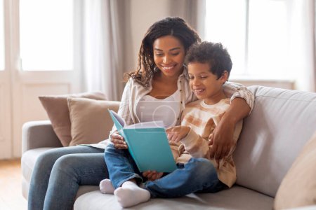 Photo for Smiling black mother and her preteen son enjoying reading book together at home, happy african american family of two sitting on comfortable sofa, sharing quiet educational moment in living room - Royalty Free Image