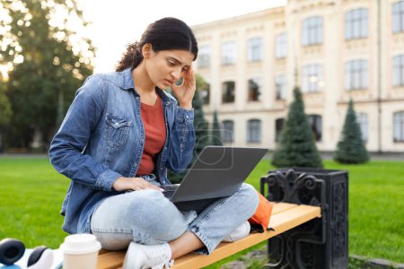 Téléchargez les photos : Jeune étudiante indienne débordée se préparant aux examens, étudiant à l'extérieur, assise sur le canapé du campus universitaire, regardant l'écran d'ordinateur portable et touchant sa tête, copiant l'espace - en image libre de droit