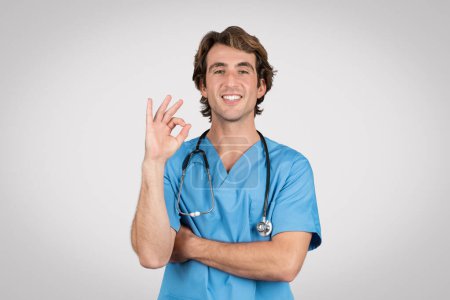 Lächelnde männliche Krankenschwester in blauem Peeling, die ein Okay-Zeichen zeigt, das die Zufriedenheit mit Gesundheitsleistungen oder positiven Patientenergebnissen repräsentiert und Vertrauen und Sicherheit in die medizinische Versorgung vermittelt