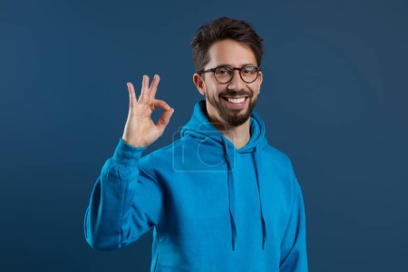 Photo for Happy Young Man Wearing Blue Hoodie Showing Ok Gesture At Camera, Cheerful Guy In Eyeglasses Gesturing Sign Of Approval, Saying Im Fine While Standing Against Dark Studio Background, Copy Space - Royalty Free Image