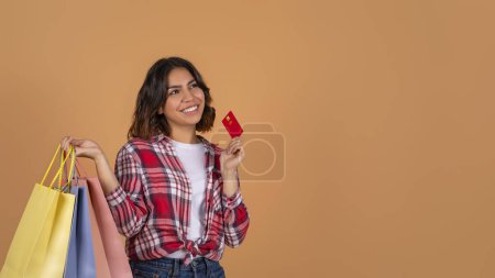 Easy shopping, cashback. Curious young middle eastern woman with purchases colorful paper bags and red plastic bank credit card looking at copy space, beige studio background, panorama