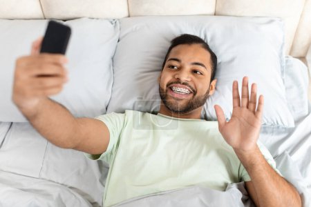 Téléchargez les photos : Vue du dessus du beau barbu afro-américain portant un pyjama couché au lit, avoir le chat vidéo du matin avec la petite amie après le réveil, agitant à l'écran du téléphone portable - en image libre de droit