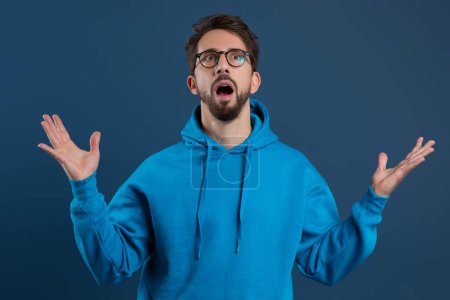 Shocked young man with beard and glasses raising hands in amazement, funny guy wearing blue hoodie with surprised face expression standing against dark studio background, symbolizing unexpectedness