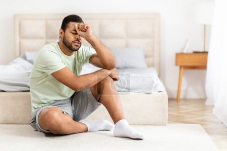 Photo for Upset young black man in pajamas sitting on floor by bed at home, thinking about something, looking for solution while 2024 crisis, leaning on his hand, panorama with copy space - Royalty Free Image