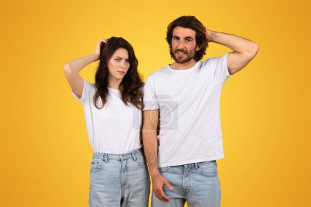 Téléchargez les photos : Jeune couple européen perplexe en t-shirts et jeans blancs, se grattant la tête avec des expressions perplexes, debout sur un fond jaune vif, incarnant confusion ou curiosité - en image libre de droit