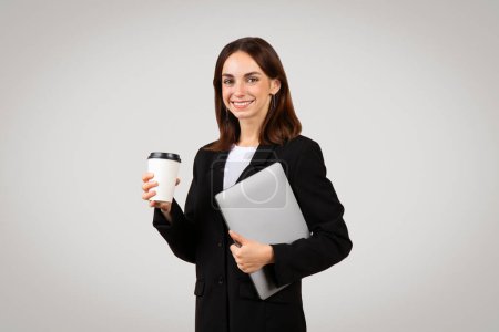 Foto de Sonriente milenaria mujer caucásica profesional vestida con traje negro sosteniendo una taza de café para llevar en una mano y una computadora portátil de plata en la otra, lista para el trabajo, aislada sobre fondo gris, estudio - Imagen libre de derechos