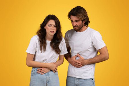 Photo for Concerned european man and woman with pained expressions holding their stomachs, possibly indicating sickness or hunger, dressed in simple white t-shirts against a yellow backdrop - Royalty Free Image