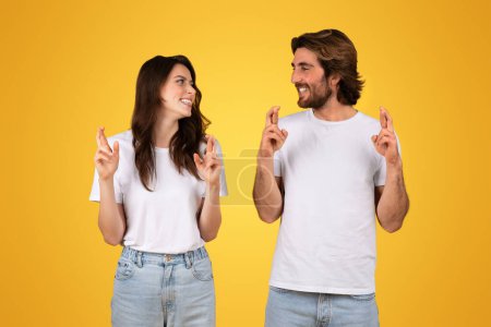 Téléchargez les photos : Optimiste souriant jeune couple caucasien en t-shirts blancs croisant les doigts pour la bonne chance, se regardant avec un sourire plein d'espoir sur un fond jaune, studio - en image libre de droit