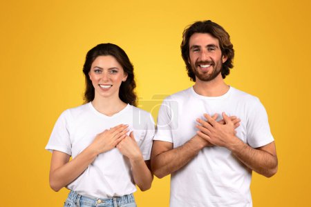 Photo for Grateful and happy millennial caucasian couple each holding hands on heart, with sincere smiles, wearing white t-shirts and standing against a cheerful yellow background, studio - Royalty Free Image