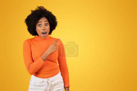 Téléchargez les photos : Choquée jolie femme afro-américaine millénaire aux cheveux bouclés pointant vers le côté, bouche ouverte dans la surprise, portant un col roulé orange, sur un fond jaune, studio - en image libre de droit