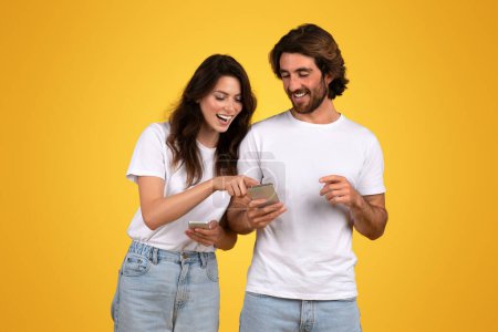 Foto de Pareja joven europea comprometida y feliz compartiendo contenido en un teléfono inteligente, riendo y apuntando a la pantalla, tanto con camisas blancas como vaqueros, sobre un fondo amarillo vibrante - Imagen libre de derechos