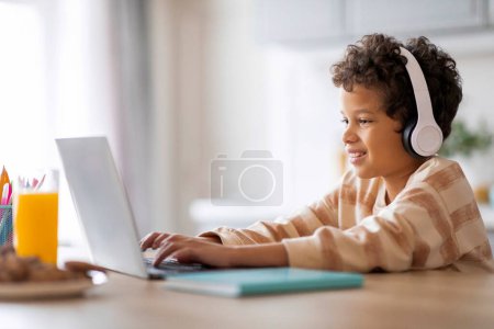 Téléchargez les photos : E-Learning. heureux écolier noir étudiant en ligne avec ordinateur portable à la maison, afro-américain mâle portant des écouteurs assis au bureau et dactylographier sur ordinateur, ayant classe à distance ou faire des devoirs - en image libre de droit