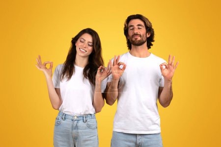 Photo for Contented european man and woman with closed eyes making OK signs, signaling approval and satisfaction in comfortable white t-shirts and jeans against a sunny yellow background - Royalty Free Image