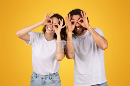 Photo for Playful european couple making funny glasses with fingers over eyes, laughing in matching white t-shirts and blue jeans, against a bold yellow background full of fun, studio - Royalty Free Image