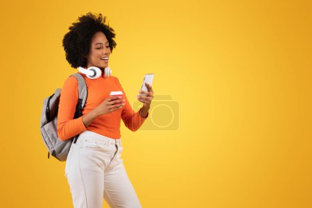 Photo for Cheerful young African American woman curly wearing an orange top smiles while looking at her smartphone, headphones around neck, holding a coffee cup, with a backpack on a yellow background - Royalty Free Image