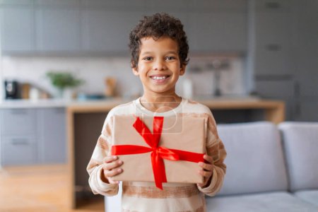 Téléchargez les photos : Joyeux petit garçon noir tenant fièrement un cadeau enveloppé avec ruban rouge vif, mignon enfant mâle préadolescent souriant à la caméra, prêt à offrir un cadeau, debout dans le salon moderne et bien éclairé intérieur, espace de copie - en image libre de droit