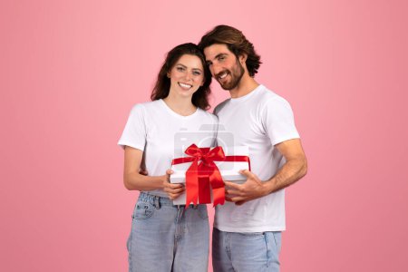 Photo for Smiling caucasian young couple in casual white t-shirts sharing a special moment, with a man giving a white gift box with a vibrant red ribbon to his partner on a pink background. - Royalty Free Image