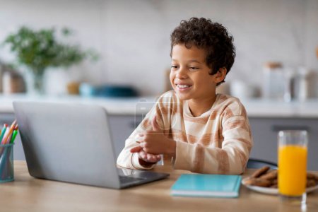 Foto de Niño negro feliz interactuando con el ordenador portátil mientras está sentado en la mesa en la cocina, con un vaso de jugo de naranja y galletas cerca, niño masculino disfrutando de un comienzo alegre de su día con el aprendizaje en línea o el ocio - Imagen libre de derechos