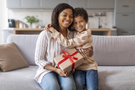 Photo for Cheerful black mother and her preteen son sharing warm embrace and holding gift with red bow, happy young mom getting gift for birthday or mothers day from her loving male kid, copy space - Royalty Free Image