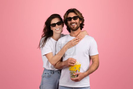 Photo for Cheerful european couple in casual wear and 3D glasses smiling and sharing popcorn, enjoying a fun moment together on a pink background, evoking a cinema date vibe, studio - Royalty Free Image