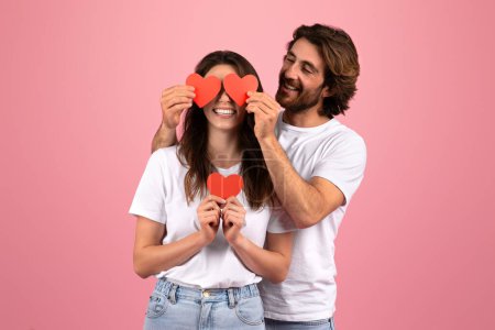 Téléchargez les photos : Joyeux couple caucasien couvre ludique leurs yeux avec des coeurs de papier rouge, partageant un moment léger ensemble, avec la femme tenant un troisième coeur contre sa poitrine, sur une toile de fond rose - en image libre de droit