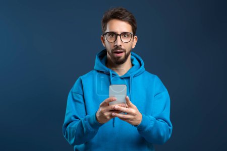 Photo for Mobile Offer. Surprised young man holding smartphone and looking at camera with excitement, millennial guy emotionally reacting to online news, standing against blue studio background, copy space - Royalty Free Image