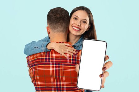 Photo for Dating Application. Happy Young Couple Hugging, Woman Showing Large Smartphone With Blank Screen, Recommending New App Or Mobile Service Over Blue Studio Background. Mockup, Collage - Royalty Free Image