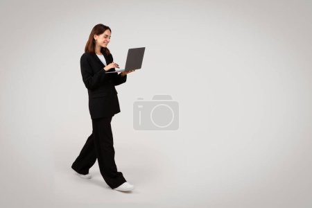 Téléchargez les photos : Femme d'affaires caucasienne souriante du millénaire en mouvement, marchant en regardant attentivement un ordinateur portable gris qu'elle tient, représentant la productivité et la mobilité dans un environnement de travail - en image libre de droit