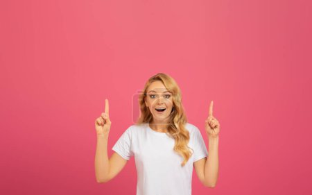 Photo for Portrait of joyful beautiful blonde lady pointing fingers up with excited expression, over pink background in studio, showcasing free space with upward gesture with both hands - Royalty Free Image