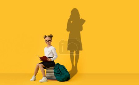 Téléchargez les photos : Une jeune fille avec des lunettes et des cheveux en petits pains s'assoit sur une pile de livres avec un sac à dos vert, lisant intensément, son ombre coulant une silhouette professionnelle aspirante contre un mur jaune - en image libre de droit