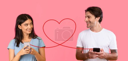 Photo for A beaming man and smiling woman, both in white t-shirts, are engaged with smartphones, connected by large red heart symbol on a pink background, portraying a cheerful online romantic interaction - Royalty Free Image
