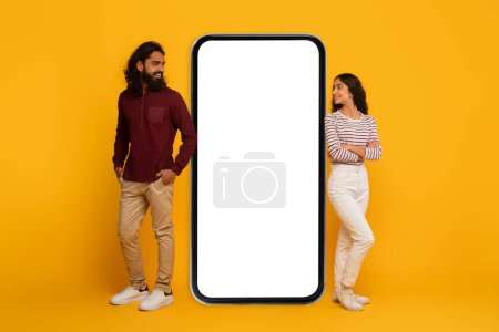 Téléchargez les photos : Homme et femme de chaque côté d'un grand écran de smartphone vierge, simulant l'interaction sur un fond jaune - en image libre de droit