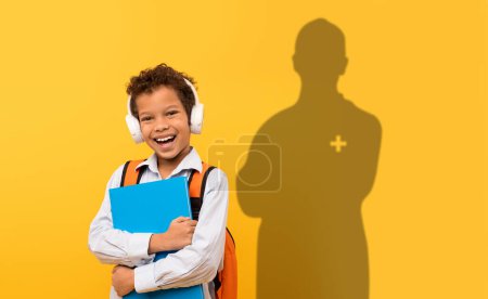 Foto de Un niño radiante con auriculares y una mochila naranja abraza su carpeta azul, su sombra con una cruz médica que sugiere un futuro en la atención médica, en un contexto amarillo soleado - Imagen libre de derechos