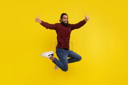 Photo for Carefree happy bearded millennial indian guy jumping up in the air and gesturing showing thumb ups isolated on yellow studio background, recommending nice good or service, full length - Royalty Free Image