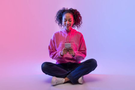 Cheerful black curly-haired lady in pink hoodie sitting cross-legged, engrossed in her smartphone on soft neon pink and blue glow neon backdrop