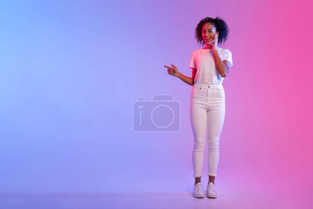 Téléchargez les photos : Joyeux dame noire avec les cheveux bouclés pointant vers le côté à l'espace libre et souriant, présentant la publicité sur fond dégradé bleu néon et rose, pleine longueur - en image libre de droit