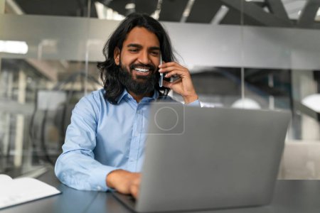 Homme barbu dans une chemise bleue sourit tout en ayant une conversation joyeuse sur son smartphone dans le bureau