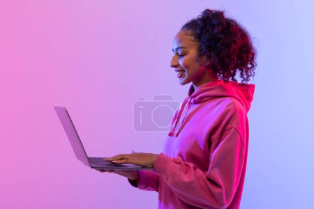 Téléchargez les photos : Radieuse jeune femme noire aux cheveux bouclés tenant un ordinateur portable, souriante en regardant l'écran, vêtue d'un sweat à capuche rose sur fond de néon rose et bleu lumineux - en image libre de droit