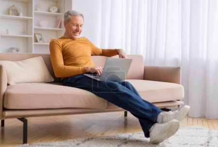 A senior man sits back on a sofa with a laptop on his lap, portraying relaxed focus in a home environment