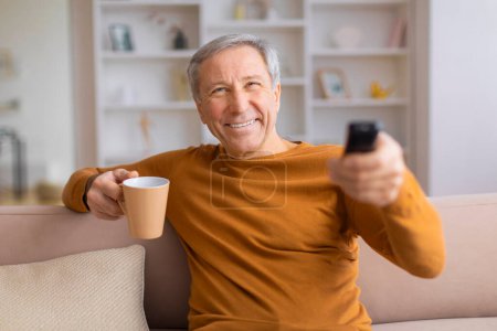 Téléchargez les photos : Un homme âgé en tenue décontractée tient une tasse à café et une télécommande, souriant en regardant vers la caméra - en image libre de droit