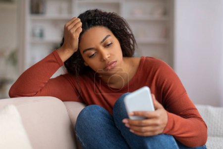 Photo for Young black woman feeling concerned while looking at her smartphone at home, upset african american female reading unpleasant message or waiting call, sitting on couch in living room - Royalty Free Image