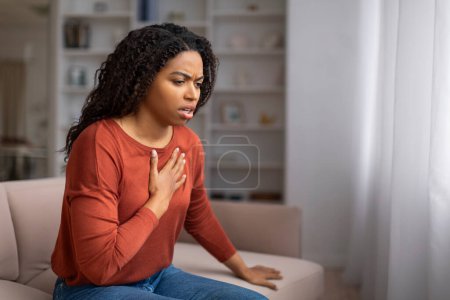 Téléchargez les photos : Inquiète jeune femme noire souffrant de douleur dans la poitrine alors qu'elle était assise sur le canapé, femme afro-américaine éprouvant de l'inconfort ou de la détresse, ayant des symptômes de crise cardiaque, se sentant mal à l'aise à la maison - en image libre de droit