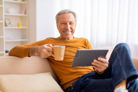 A smiling senior holds a coffee mug while using a tablet, exuding a sense of relaxation and enjoyment in his home