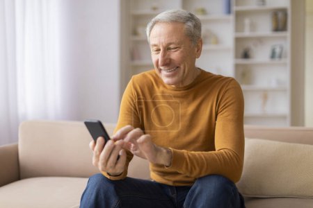 Joyful senior citizen having a fun time using his smartphone, seated on a sofa in a bright room